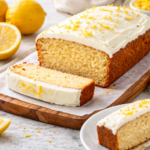 Keto lemon cake loaf with cream cheese frosting on a wooden board, served with sliced pieces and fresh lemons in a bright kitchen setting.